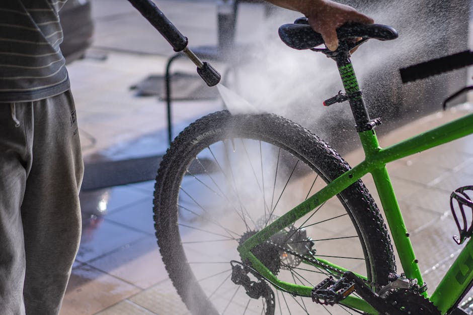 A man cleans a green bicycle with a hose, focusing on the wheel and frame outdoors.