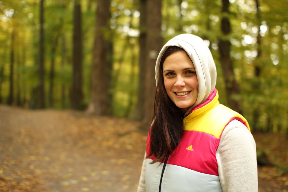 A brunette woman in a hoodie and vest smiles while hiking in a forest in Slovakia.