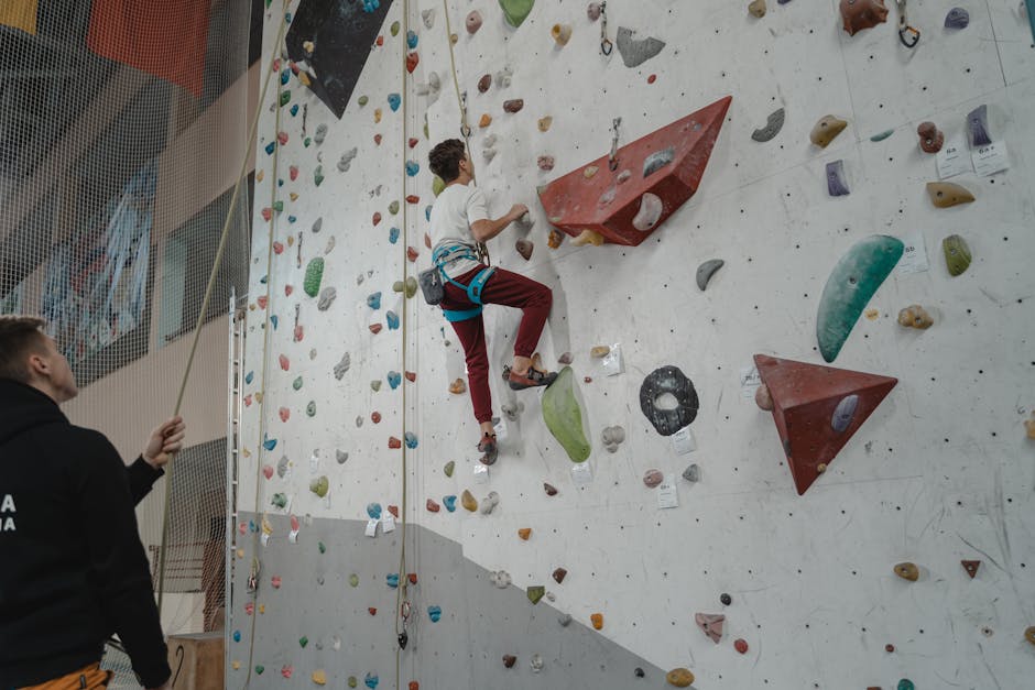 A young man practices climbing on an indoor rock wall, supervised by a trainer.