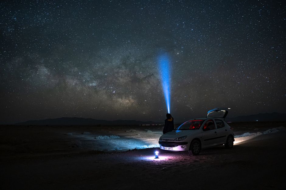 A person stands by a car under the Milky Way, using a flashlight to explore the starry night sky.