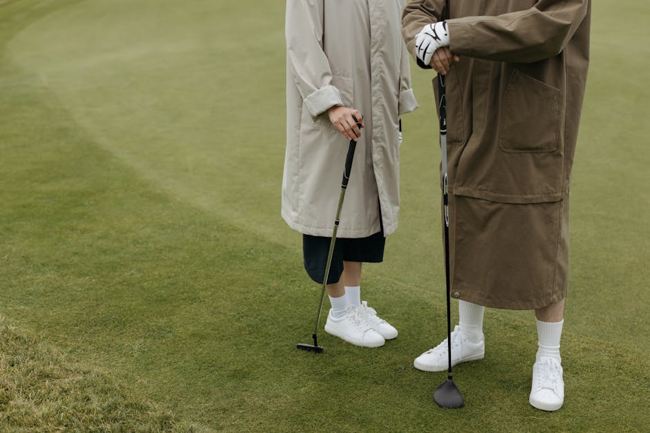 Two people in coats standing on a golf course holding clubs, ready to play.