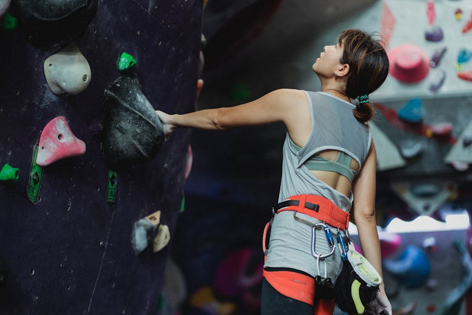 Side view of tired young Asian female athlete with chalked hands in sportswear and harness grabbing grip and looking up before climbing wall in gym